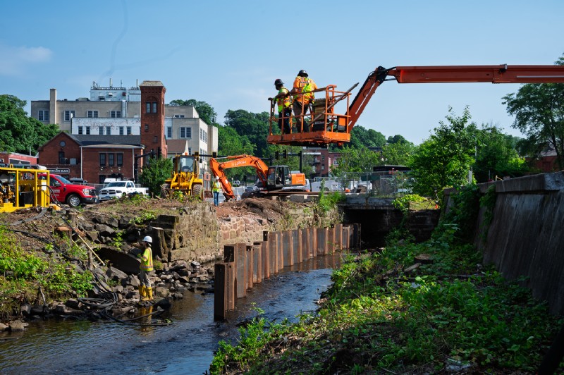 Workers on a crane oversee the bridge replacement project, with an excavator working in the background.