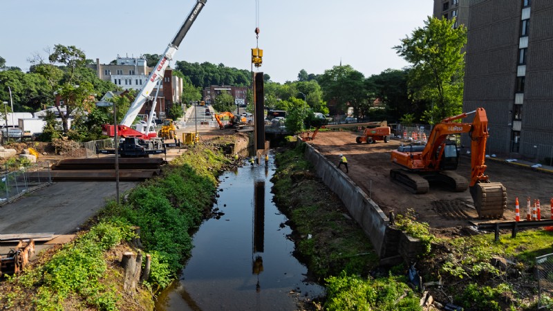 A long view of Harbor Brook as a crane installs sheet piles, with an excavator moving earth on the right bank.