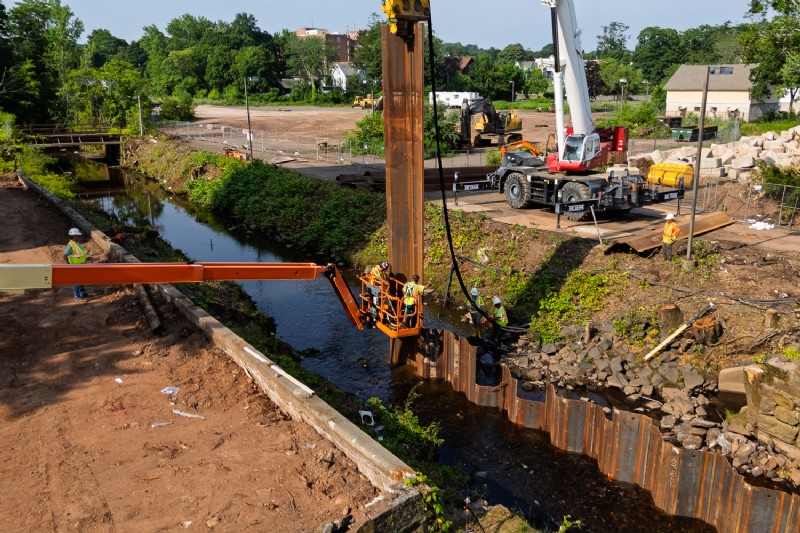 Cranes being utilized on both banks of Harbor Brook to install steel sheet piles.