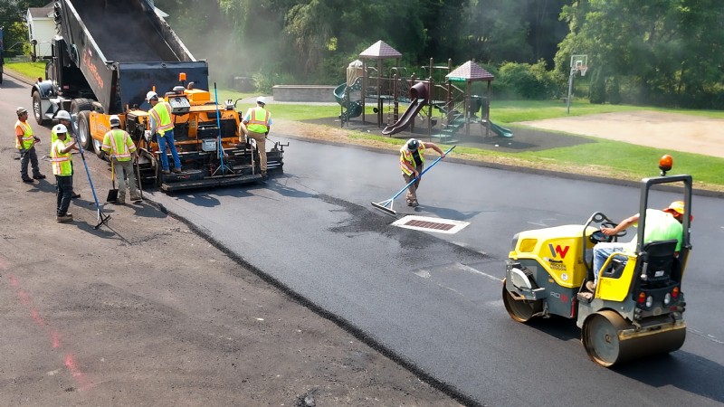 Crews using a roller and other heavy equipment to pave the parking lot, with a children’s playground in the background.