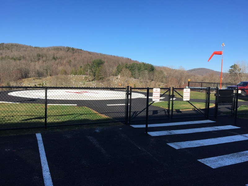 A newly installed helicopter landing pad, with fresh asphalt and white markings, surrounded by a fence.