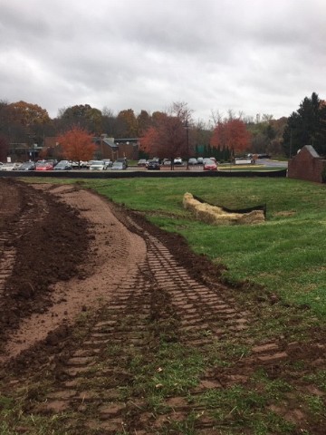 Tracks from heavy machinery cut across a field, with a parking lot in the background.