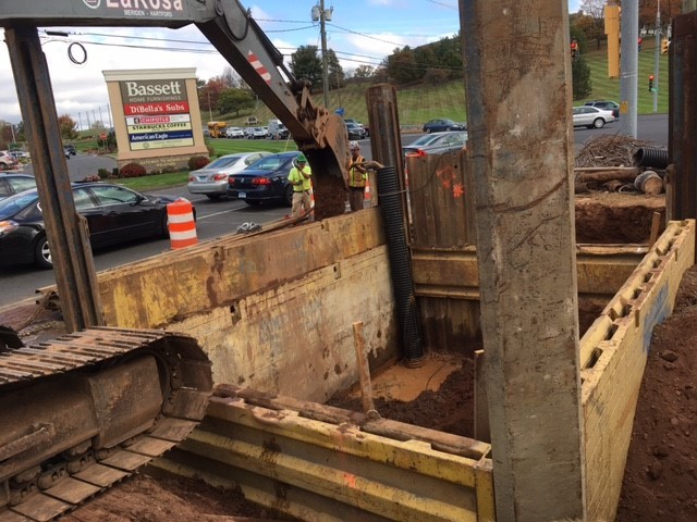 A LaRosa excavator working near the trench, with signs for a shopping center in the background.