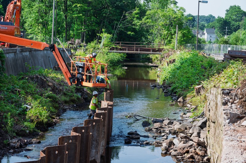 Workers on a crane are assisted by one worker standing next to steel sheet piles in Harbor Brook.